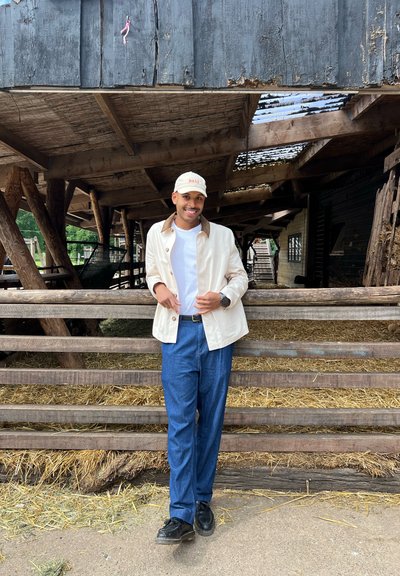 Una persona con una chaqueta color crema y cuello marrón, camisa blanca y pantalones azules está de pie junto a una cerca de madera, con heno de fondo.