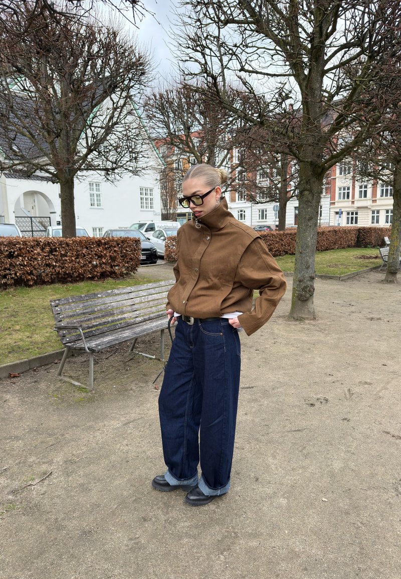 Personne avec des lunettes portant une veste marron et un large jean bleu, debout près d'un banc vide dans un parc avec des arbres sans feuilles et des bâtiments en arrière-plan.