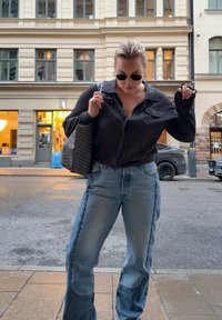 Femme portant des lunettes de soleil, un chemisier sombre et un jean bicolore, debout dans une rue de la ville, tenant un sac fourre-tout sur l'épaule.
