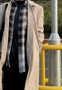 Trench-coat léger beige porté sur une chemise noire à manches longues et une chemise à carreaux noir et blanc. Pantalon noir. Rampe jaune en arrière-plan.