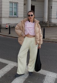 Giacca imbottita di un beige chiaro, top corto rosa e pantaloni larghi gialli chiari. Occhiali da sole neri e una borsa tote nera. Sfondi di un attraversamento pedonale.