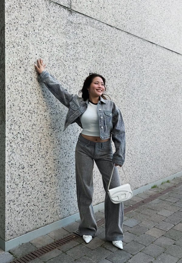 Ung kvinde iført grå denimjakke og bukser, hvid crop top og hvide hæle, smilende mens hun læner sig op ad en plettet betonvæg.