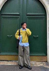Gul strikket sweater, blå skjorte med krave, beige vidbenede bukser, holder en lyseblå vandflaske, står foran en grøn dør.