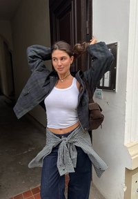 Črna karirana jakna, bela majica brez rokavov, modri denim hlače in zavezana karirana srajca. Rjava torba visoko prevrta čez ramo. Ohlapno mestno okolje.