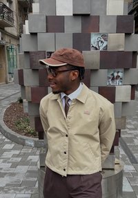 Μπεζ ελαφρύ μπουφάν με μαύρα κουμπιά, φορεμένο πάνω από λευκό πουκάμισο και καφέ γραβάτα. Καφέ καπέλο ολοκληρώνει την εμφάνιση σε μια υφασμάτινη φόντο.