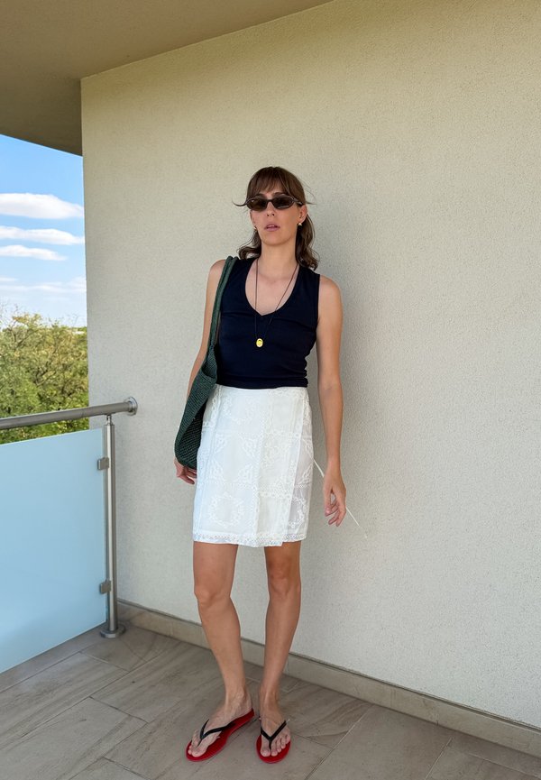 Jonge vrouw met een zwart tanktop, witte rok, rode slippers, zonnebril en een groene schoudertas, staand op een balkon.