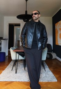 Svart skinnjakke med glidelåslukking, brukt over en svart genser. Sorte solbriller og vide svarte bukser skiller seg ut mot en lys bakgrunn.