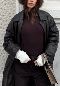 Femme portant un manteau en cuir noir, un haut foncé, des gants sans doigts blancs, un chapeau marron en fourrure, tenant un sac à main beige à motifs contre un mur texturé.