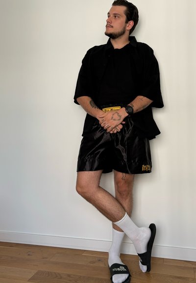Camisa negra de manga corta, pantalones cortos de satén negros con logo amarillo, calcetines blancos y sandalias negras de deslizar con logo blanco sobre un suelo de madera.