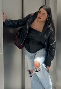 Giacca di pelle nera sopra un top senza spalline nero, abbinata a jeans chiari strappati. In piedi in un ascensore con una borsa bordeaux.