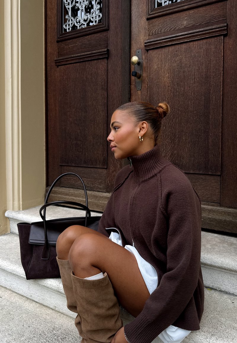 Mujer con el cabello recogido en un moño, vestida con un cuello de tortuga marrón, una falda blanca y botas color beige, sentada en unos escalones junto a una puerta de madera oscura con un gran bolso negro.