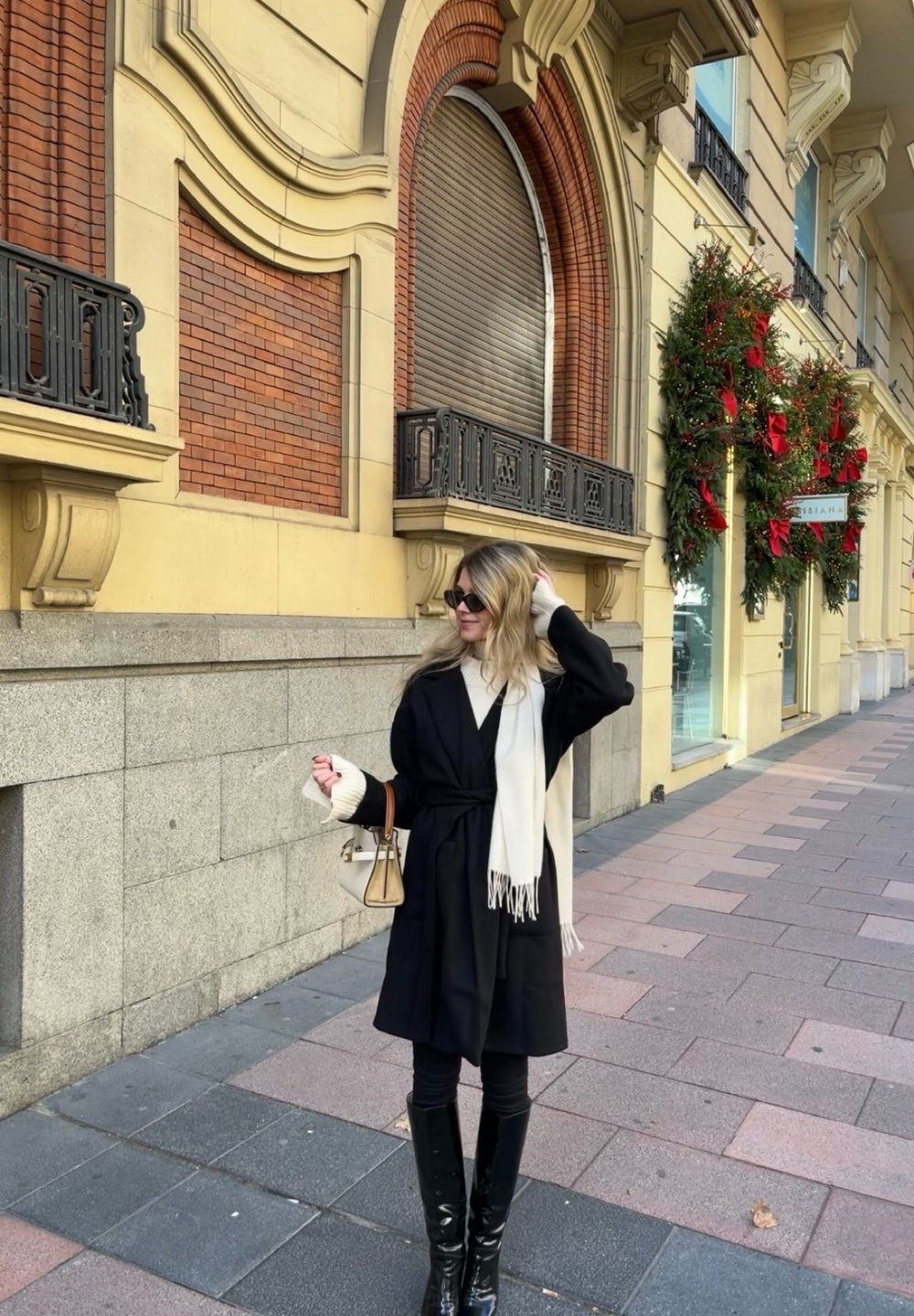 Μαύρο παλτό με λευκό μαντήλι, στενά μαύρα παντελόνια και γυαλιστερές μαύρες μπότες μέχρι το γόνατο, στέκεται σε μια ψηφιδωτή οδό κοντά σε διακοσμητική βλάστηση.