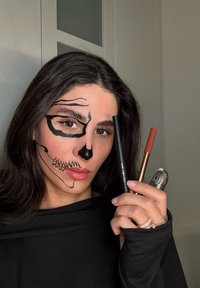 Gezicht geschilderd met zwarte ontwerpen die op een schedel lijken, met make-uptools: een zwarte eyelinerpotlood, een rood potlood en een doorzichtig voorwerp, tegen een neutrale achtergrond.