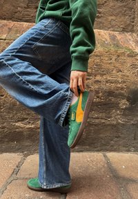 Green and orange Puma trainers made from suede and mesh, paired with blue denim flared jeans. The individual lifts their left foot to showcase the shoe details.