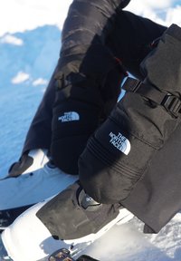 Nærbillede af sorte The North Face-snefætrebukser og handsker, båret af en person, der justerer hvide snowboardstøvler i sneen.