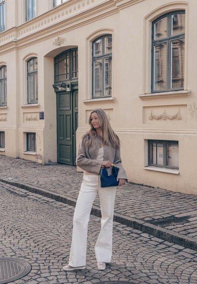 Mujer con cabello largo está de pie en una calle empedrada sosteniendo un bolso azul, frente a un edificio histórico beige con puerta verde y grandes ventanas.