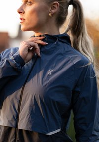 Tumesinine tuulejope kerge kangaga, must tõmblukk ja helkivad hallid detailid. Omadusteks on kõrge krae ja elastsed varrukaservad.