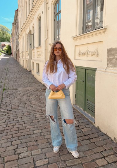Mujer de pelo largo con gafas de sol, camisa blanca, vaqueros rotos y zapatillas blancas sostiene un bolso amarillo en una calle adoquinada junto a edificios antiguos.