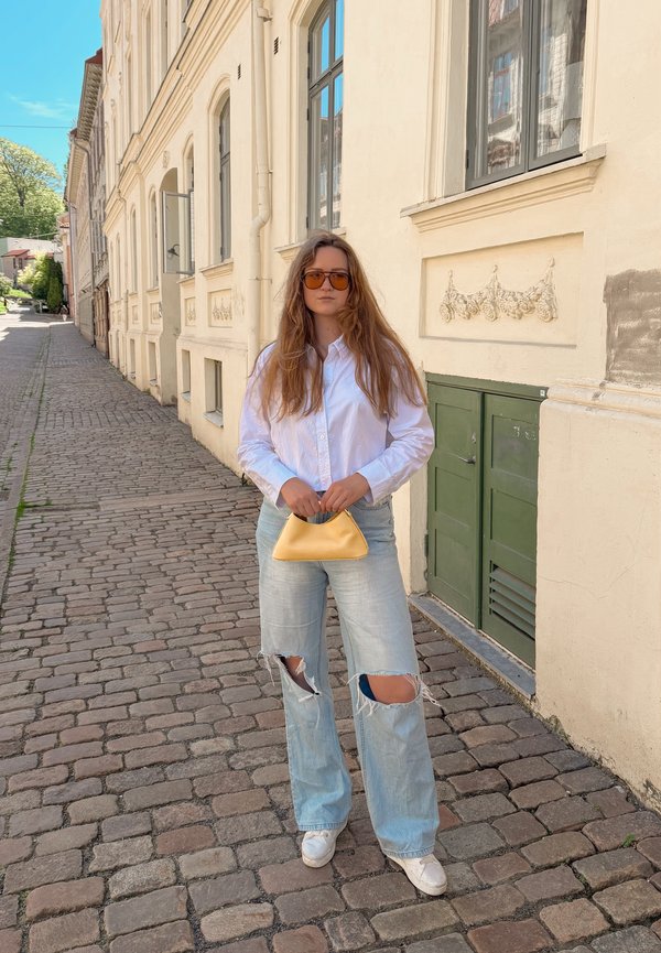 Frau mit langen Haaren, die eine Sonnenbrille, ein weißes Hemd, zerrissene Jeans und weiße Sneaker trägt, hält eine gelbe Handtasche auf einer Kopfsteinpflasterstraße neben alten Gebäuden.