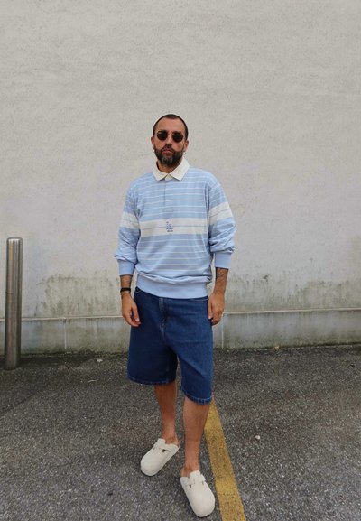 Pullover a rayas azul claro con cuello blanco, combinado con pantalones cortos de denim. Zuecos beige con hebilla completan el conjunto. El fondo es una pared lisa.