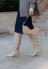 Stivali beige al ginocchio con punta affusolata e tacco a stiletto, abbinati a una borsa beige chiaro con un design curvo.