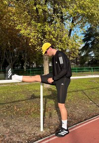 Deportista con camiseta negra de manga larga con tres rayas blancas, pantalones cortos negros, gorra amarilla y zapatos negros estirando la pierna contra una barandilla blanca.