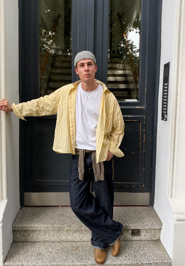 Jeune homme portant une chemise rayée jaune, un t-shirt blanc et une casquette à carreaux, appuyé contre un cadre de porte sombre avec des escaliers visibles à travers des panneaux en verre.