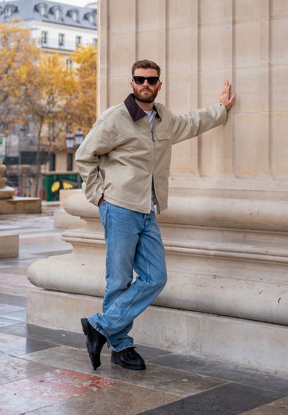 Veste beige à fermeture éclair avec col marron, associée à un jean bleu clair et des chaussures noires. Mur en pierre texturé en arrière-plan.