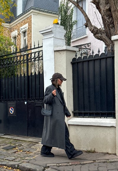 Abrigo gris de doble botonadura llevado sobre un conjunto negro, combinado con una gorra negra y un bolso pequeño, caminando por un camino de piedra al lado de una entrada cerrada.