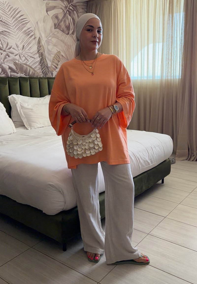 Mulher usando lenço leve na cabeça, blusa laranja de manga comprida e calças brancas, segurando uma pequena mala branca num quarto com uma cama verde.