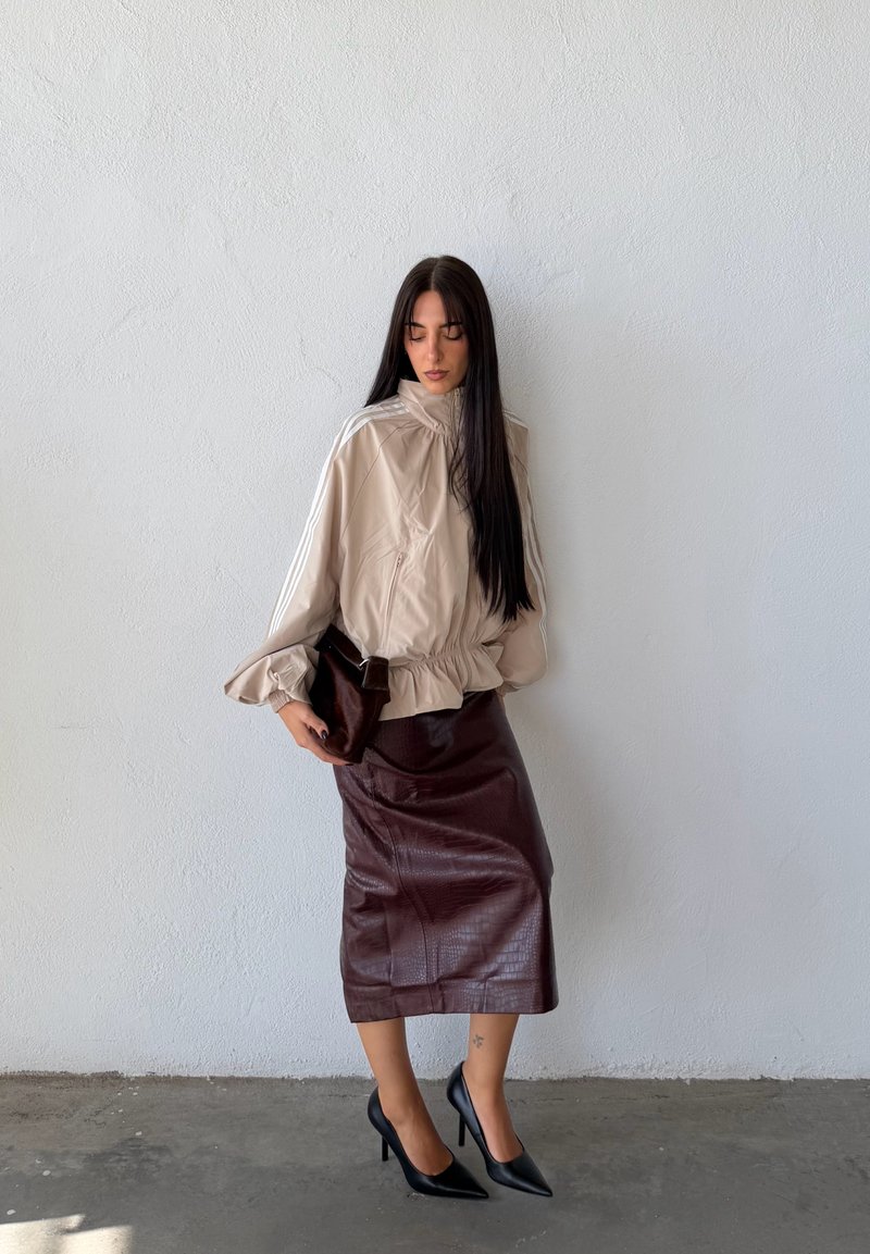 Une femme pose devant un mur blanc. Elle porte une veste beige zippée, une jupe en cuir bordeaux et des talons noirs, avec un sac porté à l’épaule de couleur foncée.