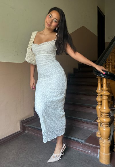 Vestido blanco de encaje con forma ajustada, que presenta un tirante y tejido texturizado. Los accesorios incluyen tacones deslizantes beige y un bolso a juego.