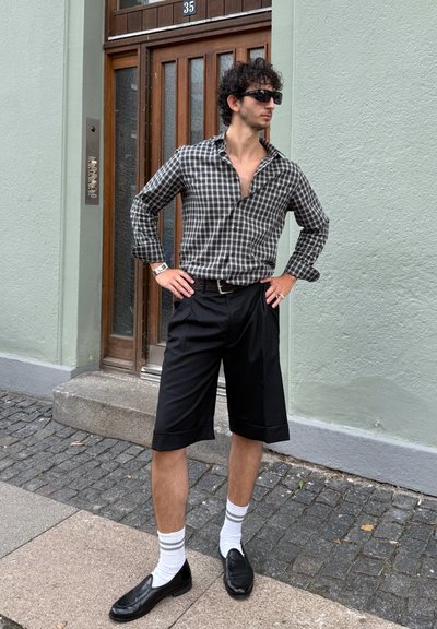 Camisa a cuadros en blanco y negro, pantalones cortos negros hasta las rodillas, mocasines negros y calcetines blancos con rayas grises, de pie cerca de una puerta de madera.