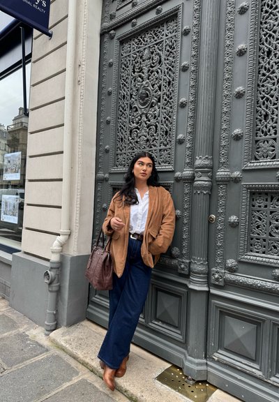 Chaqueta oversized de ante marrón, camisa blanca de botones, pantalones de mezclilla oscuros de pierna ancha, bolso de cuero marrón y botas de tobillo, posando frente a una puerta decorativa gris.