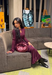 Γυναίκα με μπορντό φόρεμα και μαύρες μπότες καθισμένη σε γκρι καναπέ με μπορντό τσάντα, τρεις πολύχρωμοι πίνακες ζωγραφικής τοποθετημένοι σε καβαλέτα πίσω της.