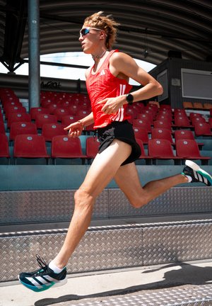 Atleta che corre sulle scale, indossando una canotta rossa senza maniche con righe, pantaloni corti neri e scarpe sportive nere con accenti verdi.