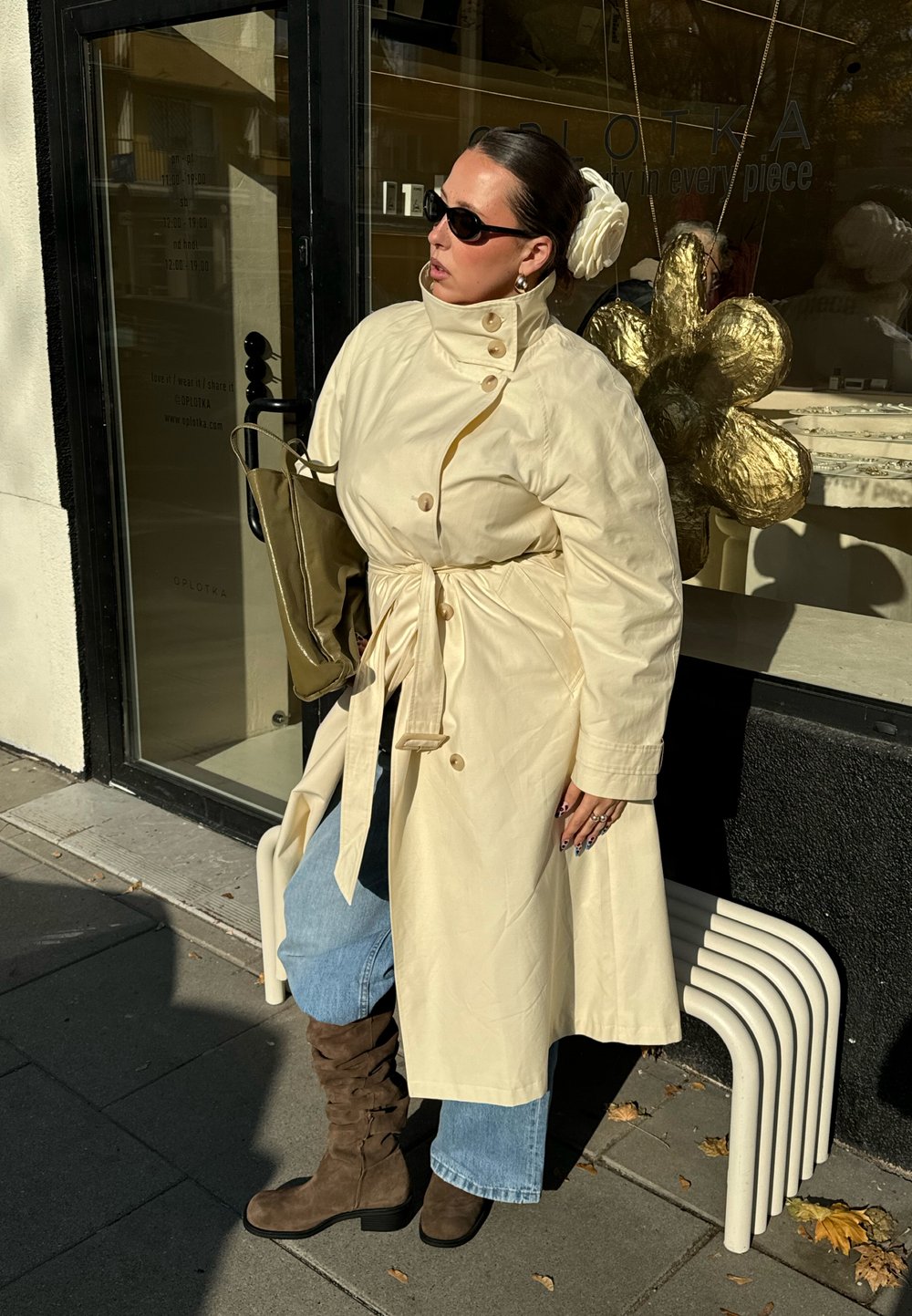 Krém színű ballonkabát gombos részletekkel és övvel, kék bőszárú farmerrel és barna, lazán álló csizmával párosítva. Zöld táska és napszemüveg.