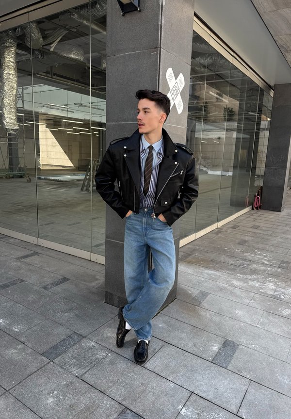 Jonge man leunt tegen een pilaar en draagt een zwarte leren jas, gestreept hemd, das, wijde blauwe jeans en zwarte schoenen op een stedelijk trottoir.