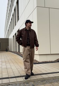 Veste marron à fermeture éclair sur un pull à motifs, pantalon large beige et chaussures noires, sur fond d'un extérieur de bâtiment moderne.