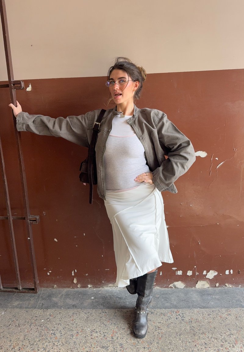 Jeune femme portant des lunettes, une veste grise, une jupe blanche et des bottes noires, s'appuie d'un bras sur des barres métalliques contre un mur marron bi-ton.