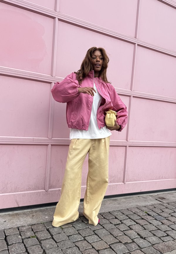 Žena u ružičastoj jakni i širokim bež hlačama drži malu žutu torbu stojeci na kaldrmisanoj ulici uz ružičasti zid.