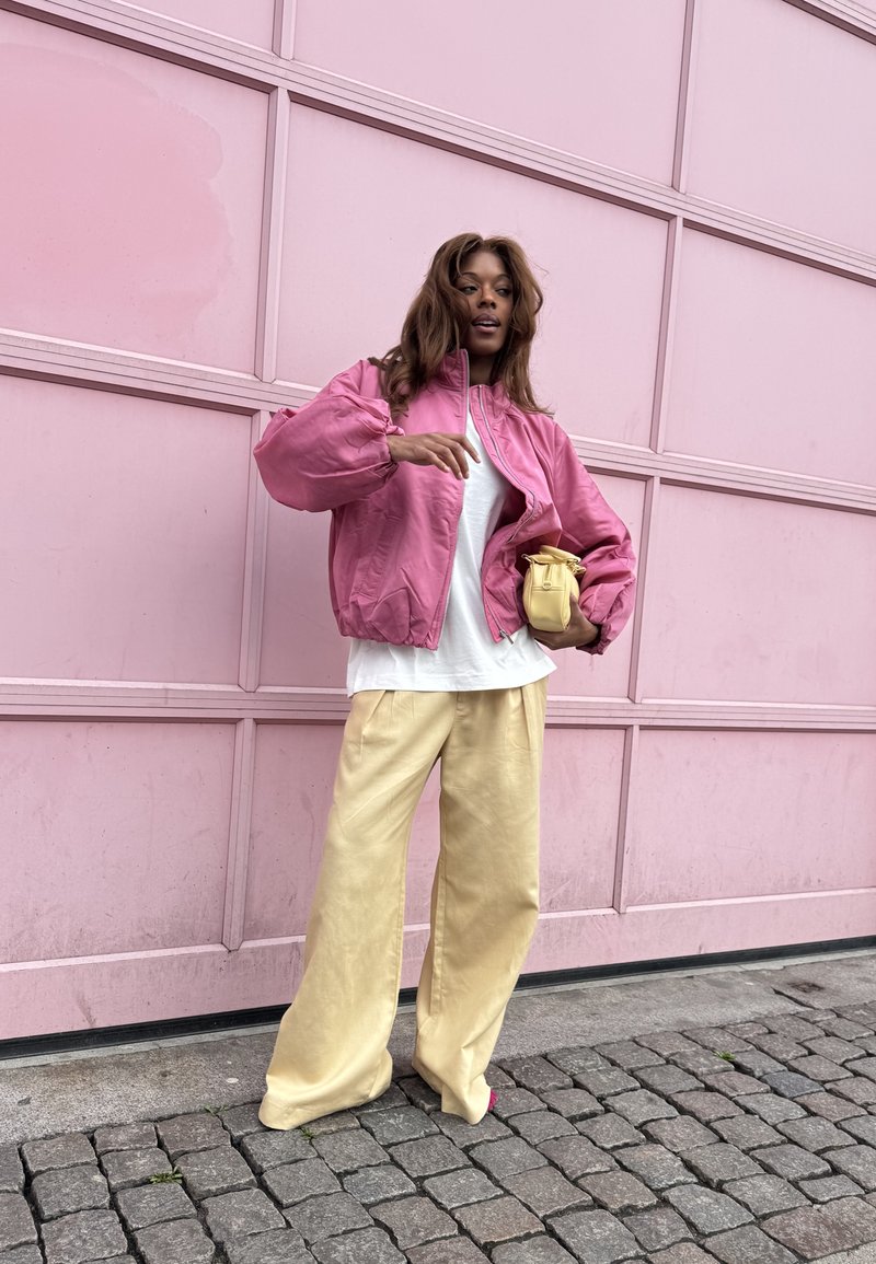 Γυναίκα με ροζ μπουφάν και φαρδιά μπεζ παντελόνια κρατά μικρή κίτρινη τσάντα και στέκεται σε πλακόστρωτο δρόμο μπροστά από ροζ τοίχο.
