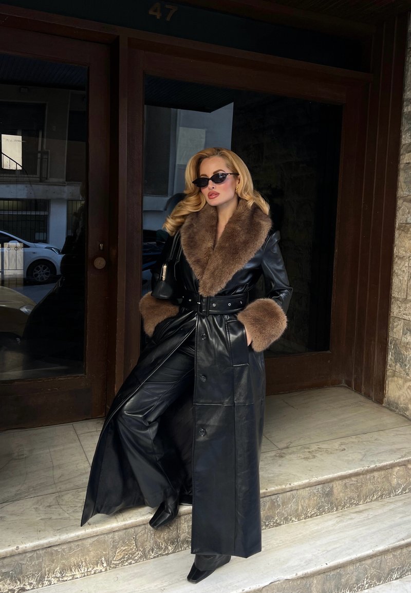 Femme aux cheveux blonds portant un manteau en cuir noir avec un col et des poignets en fourrure marron, pantalon noir, lunettes de soleil, debout sur des marches en pierre à côté d'une porte en bois.