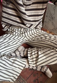 Personne assise en tailleur portant une chemise à rayures marron, un pantalon blanc avec des rayures marron, et des chaussettes beiges avec des motifs foncés sur un tapis à motifs.