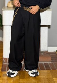 Fekete, bőszárú nadrág magas derékkal, aranylánc övvel díszítve. Vastag fehér-fekete sportcipőkkel párosítva, fapadlón.