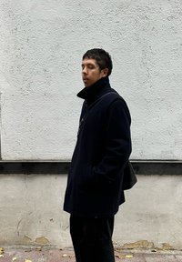 Abrigo azul marino de lana con cuello alto, usado sobre pantalones negros. Bolsa tipo mensajero negra colgada al hombro. Pared blanca lisa y texturizada al fondo.