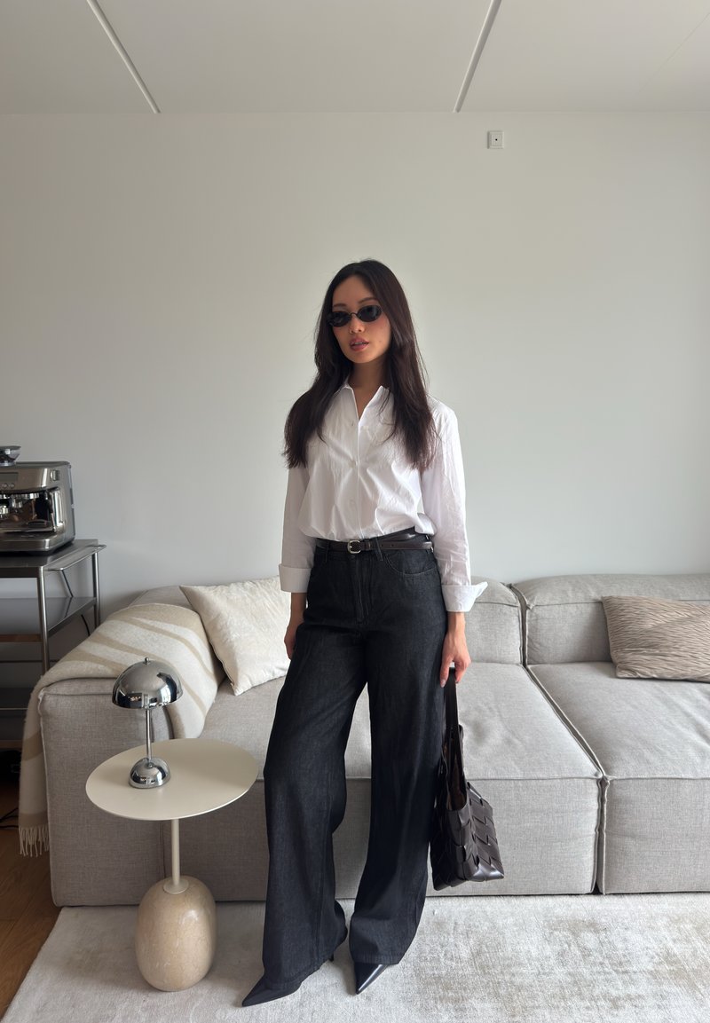 Femme portant des lunettes de soleil, une chemise blanche et un pantalon noir large, se tient dans un salon moderne tenant un sac à main noir près d’un canapé gris et d’une table ronde.