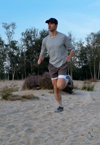 Uomo che corre su una superficie sabbiosa, indossando una maglietta grigia a maniche lunghe, shorts marroni con bordi bianchi e scarpe da corsa grigie, con un cappellino nero.