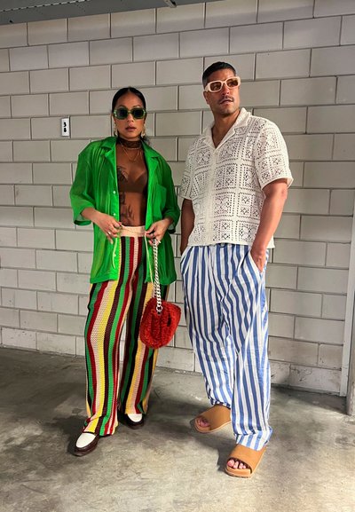 Dos individuos están de pie frente a una pared texturizada. Uno lleva una camisa verde transparente, pantalones a rayas y sostiene un bolso rojo de peluche. El otro tiene una camisa blanca con un patrón, pantalones azules a rayas y chanclas marrones.