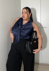 Femme portant un haut sans manches en satin bleu marine et un pantalon noir, tenant un sac à main noir, se tenant à l'intérieur devant une armoire blanche.